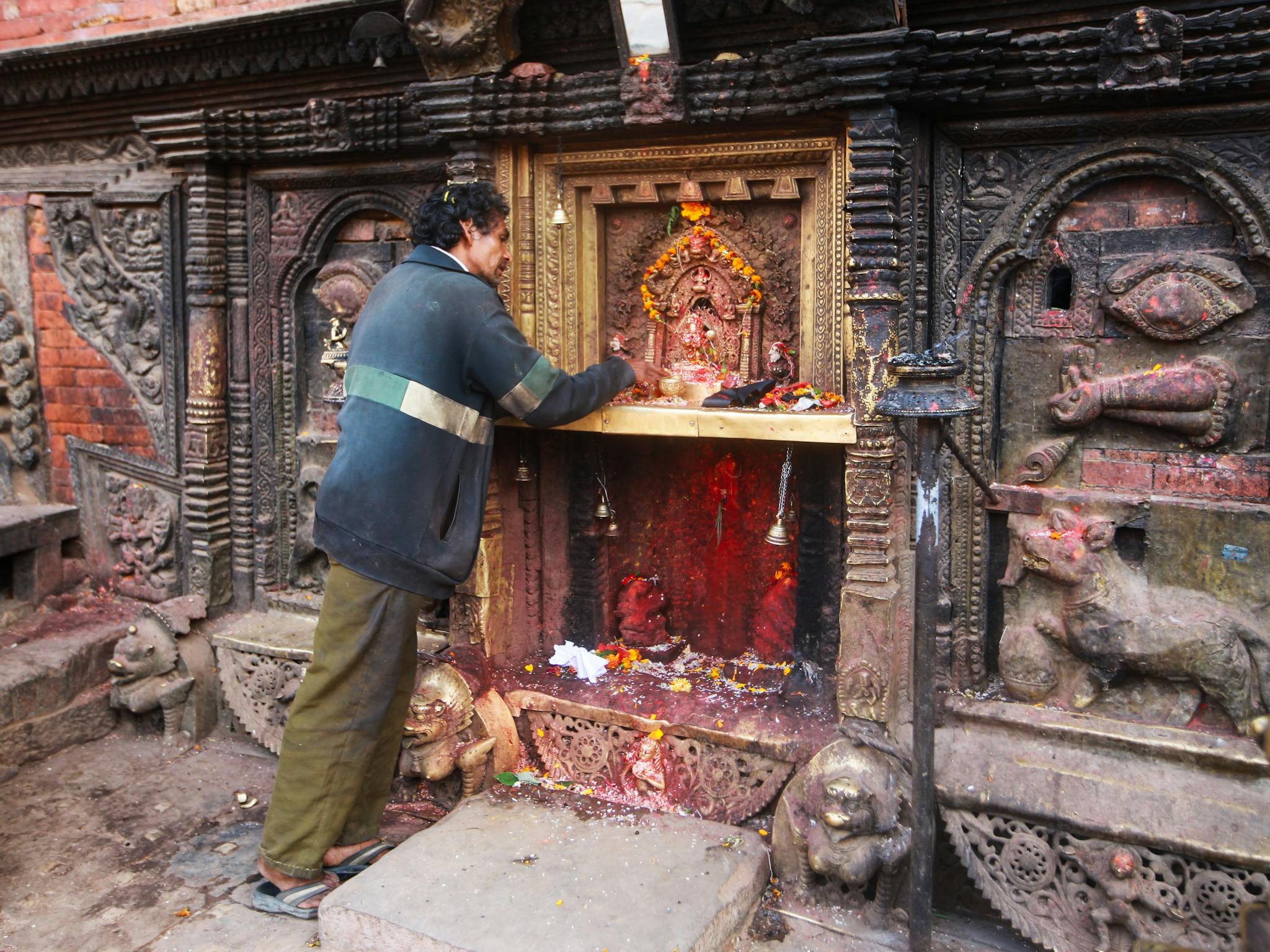 A man engages in prayer at an ornate stone shrine adorned with rich carvings and offerings.